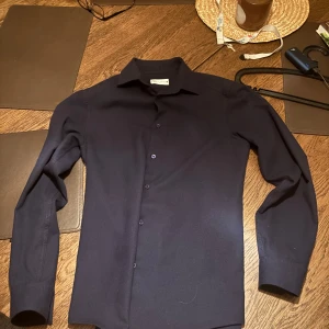 Mörkblå skjorta från  - Säljer en stilren skjortan som har klassisk krage, knäppning framtill och långa ärmar. Perfekt för dig som vill ha en enkel men snygg skjorta till garderoben. Knappt använd och perfekt för sommaren