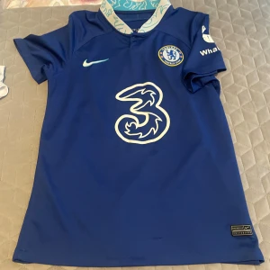 Chelsea FC hemmatröja 2022/23 S  - Säljer en blå Chelsea FC fotbollströja från Nike med klubbmärke och sponsortryck på bröstet. Tröjan har korta ärmar och klassisk V-ringning med mönstrad insida på kragen. Perfekt för fans som vill visa sitt stöd på matchdag!