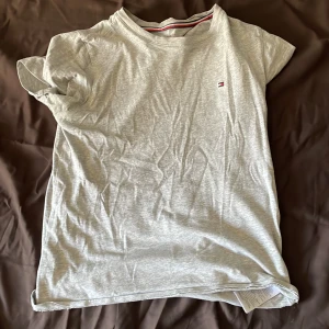 Grå t-shirt från Tommy Hilfiger - Säljer en klassisk grå t-shirt från Tommy Hilfiger med liten broderad logga på bröstet. T-shirten har rund halsringning och korta ärmar. Perfekt basplagg med diskret design och mjukt bomullsmaterial.