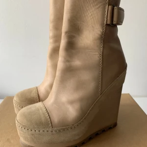 Beige kilklackade boots från Acne - Säljer ett par beige boots från Acne med kilklack och rund tå. Skorna är i skinn och mocka med grov sula och dragkedja på sidan. De har även ett spänne upptill som detalj. Perfekta för dig som vill ha både höjd och komfort.