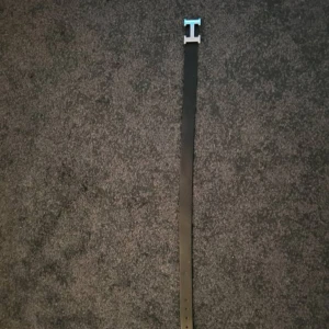 Svart bälte med silverfärgat H-spänne - Säljer ett stilrent svart bälte med ett stort, silverfärgat H-format spänne. Bältet är smalt och har en klassisk design som passar till många olika outfits. Perfekt accessoar för dig som vill ha en snygg detalj i din look.