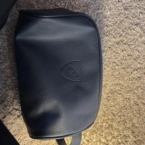 Mörkblå necessär med DIF-logga - Säljer en mörkblå necessär i skinnimitation med Djurgårdens IF:s logga präglad på framsidan. Väskan har dragkedja upptill och ett justerbart axelband med en liten randig detalj. Perfekt för att bära det viktigaste till träning eller vardag.