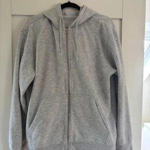 Grå hoodie med dragkedja - En klassisk grå hoodie från weekday med dragkedja framtill och huva med snören. Tröjan har två stora fickor på magen och långa ärmar. Perfekt för en avslappnad stil.