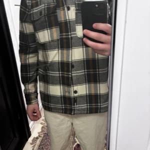 Rutig overshirt i beige och svart - Säljer en snygg overshirt med rutigt mönster i beige, svart och bruna toner. Skjortan har långa ärmar, knappar framtill och bröstficka. Perfekt att slänga över en t-shirt för en avslappnad look. Passar bäst på någon som är 178-188cm