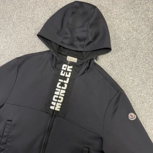 Hoodie från Moncler | ev byte - Svart vindjacka från Moncler med huva och dragkedja. Stort Moncler-tryck längs dragkedjan och klassisk logotyp på ärmen. Två fickor framtill och stilren design, perfekt för en sportig look.