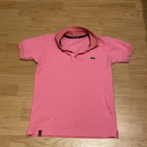Rosa pikétröja från Lacoste - Säljer en klassisk rosa pikétröja från Lacoste med korta ärmar och den ikoniska gröna krokodilloggan broderad på bröstet. Tröjan har krage och knappar framtill. Perfekt för en sportig och stilren look.