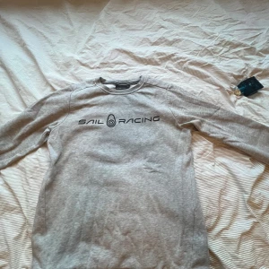 Grå sweatshirt från Sail Racing - Säljer en ljusgrå sweatshirt från Sail Racing med svart logga tryckt på bröstet. Tröjan har rund halsringning och långa ärmar. Perfekt för en avslappnad stil.