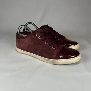 Vinröda sneakers med lackdetaljer - Snygga vinröda sneakers med ovandel i mocka och tå samt häl i blankt lackmaterial. Skorna har snörning och vit platt sula. Perfekta för dig som vill ha en stilren men ändå unik look.