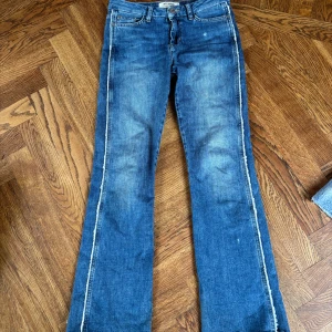 Blå bootcut low waist jeans från Mos Mosh med coola kanter - Säljer ett par blå bootcut low waist jeans från Mos Mosh med råa, fransiga kanter längs sidorna. Jeansen har klassisk femficksdesign, knapp och dragkedja framtill samt en snygg tvättad look. Så sjukt coola! Storlek 25