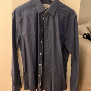 Blå mönstrad skjorta från Eton - Snygg blå skjorta från Eton med diskret rutigt mönster och klassisk krage. Skjortan har långa ärmar och vita knappar framtill samt vid ärmslut. Perfekt för dig som gillar stilrena detaljer och hög kvalitet. Den är i storlek M