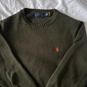 Olivgrön stickad tröja från Polo Ralph Lauren - Snygg olivgrön stickad tröja från Polo Ralph Lauren med rund halsringning och den klassiska orangea loggan broderad på bröstet. Perfekt för lager-på-lager och passar till många olika stilar.