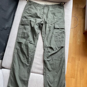 Olivgröna cargobyxor från Carhartt - Säljer ett par olivgröna cargobyxor från Carhartt med raka ben och flera praktiska fickor på sidorna. Byxorna har justerbara spännen i midjan och är tillverkade i slitstark bomull. Perfekta för dig som gillar funktionella och snygga byxor!