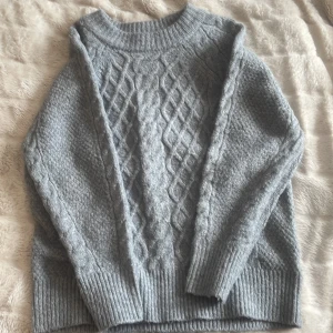 Grå stickad tröja med kabelmönster från lager 157 - Mysig grå stickad tröja från 157 i storlek XS. Tröjan har ett klassiskt kabelstickat mönster framtill och ribbade muddar vid hals, ärmslut och nederkant. Perfekt för kyliga dagar.
