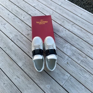 Valentino Garavani sneakers med svart detalj - Säljer ett par vita Valentino Garavani sneakers med bred svart rem över mitten. Skorna har klassisk rund tå, snörning och platt sula. Snygg och stilren design som passar till många outfits. Kommer med originalkartong och dustbag. Passar storlek 43-44. Priset kan diskuteras.