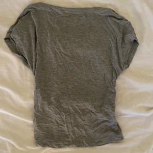 Grå t-shirt - En enkel grå t-shirt i mjukt material. T-shirten har korta ärmar och öppenrygg, perfekt för vardagsbruk. Färgen är neutral och lätt att matcha med andra plagg.
