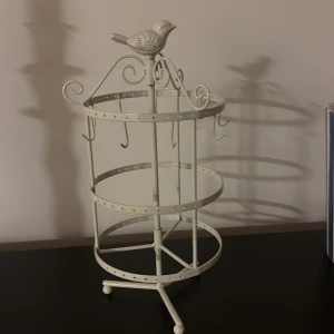 Vit smyckeshållare med fågeldekor - Säljer en vit smyckeshållare i metall med dekorativ fågel på toppen. Hållaren har flera krokar för att hänga smycken som halsband, armband eller örhängen. Perfekt för att organisera dina accessoarer på ett stilrent sätt.