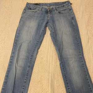 Ljusblå lågmidjade jeans - Säljer ett par ljusblå lågmidjade jeans med klassisk femficksdesign. Jeansen har raka ben och snygga detaljer på bakfickorna. Perfekta för en avslappnad stil. Ena ”hänget” har lossnat men inget man tänker på! Står ingen storlek men skulle tippa på w27!