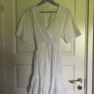 Vit omlottklänning med volang och knytband - Säljer en söt vit klänning med omlottliv, korta ärmar och volangdetaljer nertill. Klänningen har knytband i ryggen och markerad midja för en feminin siluett. Perfekt för sommarens alla tillfällen!