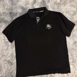 Svart pikétröja från Burberry - Klassisk svart pikétröja från Burberry med broderad logga på bröstet och rutiga detaljer vid knäppningen. Tröjan har krage och korta ärmar, tillverkad i 100% bomull för en skön känsla.