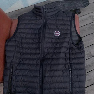 Svart dunväst från Colmar - Säljer en svart dunväst från Colmar med quiltad design och dragkedja framtill. Västen har två fickor med dragkedja och en hög krage. Perfekt för lager-på-lager och enkel att matcha med olika outfits.