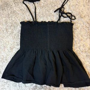 Svart smockad linnetopp med tunna axelband - Säljer en svart linnetopp med smockad överdel och tunna axelband som knyts på axeln. Toppen har en lätt utsvängd nederdel och är perfekt för varma dagar.