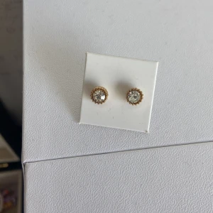 Guldpläterade örhängen med kristall från Syster P - Säljer ett par eleganta guldpläterade örhängen från Syster P med rund, glittrande kristall i mitten. Perfekta för att ge en touch av lyx till din stil. Levereras i originalpåse.