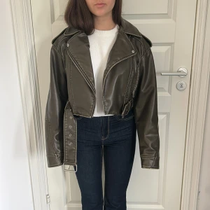 Olivgrön skinnjacka med bälte - Säljer en olivgrön skinnjacka i bikerstil med silvriga dragkedjor och bälte i midjan. Jackan har klassisk krage, långa ärmar och croppad passform. Perfekt för dig som vill ha en edgy look.