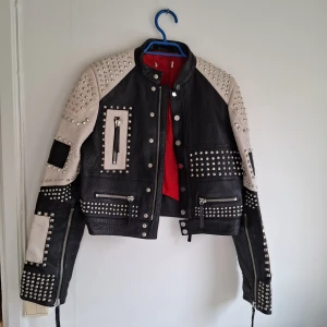Svart och beige skinnjacka med nitar - Riktigt cool svart och beige skinnjacka med massor av silvriga nitar och dragkedjor. Jackan har lång ärm, flera fickor och detaljer i både svart och beige skinn. Röd insida ger extra edge. Perfekt statement-plagg för dig som vill sticka ut.