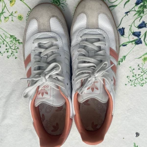 Adidas Samba i vitt och korall - Säljer ett par Adidas Samba sneakers i vitt skinn med rosa detaljer och klassiska tre ränder på sidan. Skorna har gummisula, snörning och rund tå. Perfekta för dig som gillar retrostil och vill sticka ut lite extra.