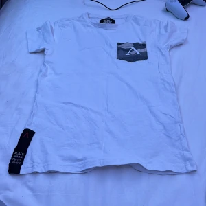 Vit t-shirt med camoflageficka från Slade Couture - Vit t-shirt från Slade Couture med camoflagemönstrad bröstficka och stort tryck med siffran 99 på ryggen. Slade-logga tryckt på baksidan. Klassisk passform och korta ärmar. Perfekt för en avslappnad streetstil.