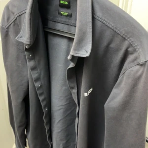 Mörkgrå skjorta från BOSS - Snygg mörkgrå skjorta från BOSS med klassisk krage och diskret logga på bröstet. Skjortan har långa ärmar och knäppning framtill. Perfekt för dig som gillar stilrena plagg med en exklusiv touch.