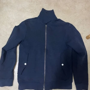 Mörkblå J.Lindeberg kofta  - Säljer en mörkblå kofta från J.Lindeberg med dragkedja och hög krage. Jackan har två fickor framtill med knappstängning och en stilren, enkel design. Perfekt för dig som gillar en clean och sportig look.