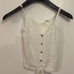 Vit linnetopp med knyt och knappar - Säljer en vit linnetopp med smala axelband, knyt längst ner och dekorativa knappar framtill. Toppen är tillverkad i 100% viskos och har en lätt och luftig känsla, perfekt för varma dagar.