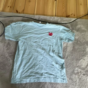 Ljusblå t-shirt från Comme des Garçons Play - Säljer en ljusblå t-shirt från Comme des Garçons Play med det klassiska röda hjärtat broderat på bröstet. T-shirten har rund hals och korta ärmar. Perfekt för en stilren och trendig look.