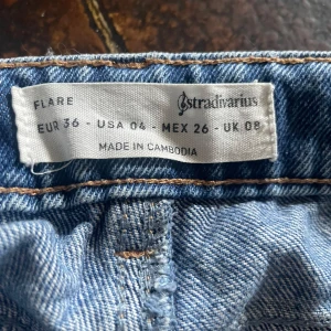 Blå flare jeans från Stradivarius - Säljer ett par blå flare jeans från Stradivarius i klassisk denim med slitningar på ena benet. Byxorna har hög midja och utsvängda ben, samt femficksmodell och gylf med dragkedja.