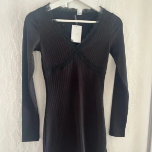 Svart ribbad klänning med spetsdetaljer - Säljer en svart ribbad klänning med v-ringning och spetsdetaljer vid halsen. Klänningen har lång ärm och figurnära passform, perfekt för dig som gillar en stilren och enkel look.