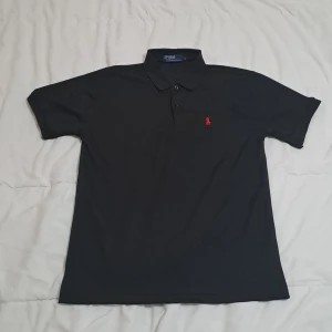 Svart pikétröja från Ralph Lauren - Klassisk svart pikétröja från Ralph Lauren med röd broderad logga på bröstet. Tröjan har korta ärmar, krage och knappar framtill. Perfekt för en stilren och sportig look.