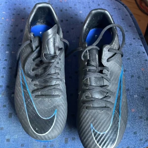Svarta fotbollsskor från Nike med blå detaljer - Säljer ett par svarta fotbollsskor från Nike med blå detaljer och stora Nike-loggor på sidorna. Skorna har snörning och dobbar i plast för spel på gräs. Snygg sportig design med blå sula och svarta ovandelar med mönster.
