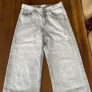 Grå jeansbyxor från Gina Tricot - Säljer ett par grå jeansbyxor från Gina Tricot i storlek 152. Byxorna har raka, vida ben och klassisk femficksdesign. De har en liten sliten detalj framtill och stängs med knapp och dragkedja.