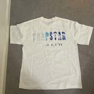 Trapstar t shirt  - Vit t-shirt från Trapstar med stort blått och svart tryck framtill och text på ryggen. Klassisk passform och rund halsringning. Perfekt för en avslappnad streetwear-look.