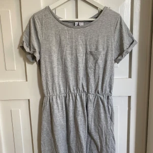 Grå t-shirtklänning från Divided - Enkel och bekväm grå t-shirtklänning från Divided med korta ärmar, rund halsringning och liten bröstficka. Klänningen har resår i midjan för en avslappnad passform och är tillverkad i mjuk bomull.