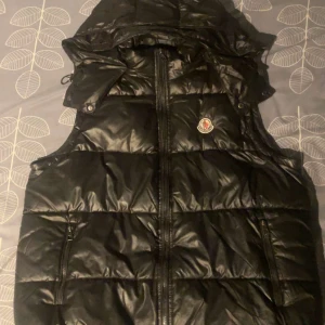 Svart dunväst från Moncler - Säljer en svart dunväst från Moncler med huva och dragkedja framtill. Västen har Moncler-logga på bröstet och en quiltad design som ger en puffig look. Perfekt för lager-på-lager-stil under kyliga dagar. Priset kan diskuteras, skriv för minsta fundering