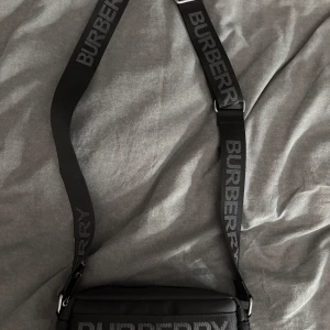 Svart axelväska från Burberry - Snygg svart axelväska från Burberry med justerbart axelband och tydlig logga. Väskan har rutigt mönster framtill och dragkedja upptill. Perfekt för att bära det viktigaste med stil.