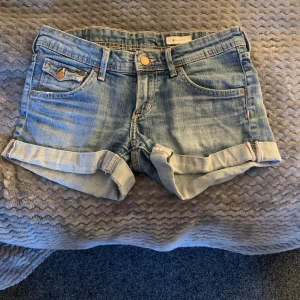 Ljusblå jeansshorts  - Säljer ett par ljusblå jeansshorts med klassisk femficksdesign och uppvikta benslut. Bakfickorna har knappar som en snygg detalj. Perfekt för varma dagar!