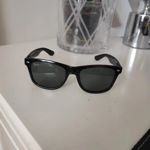 Svarta solglasögon från Ray-Ban - Klassiska svarta solglasögon från Ray-Ban med Wayfarer-form. Bågarna är i svart plast och glasen är mörka. Perfekt accessoar för soliga dagar och tidlös stil.