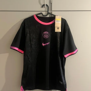 Svart PSG fotbollströja med rosa detaljer - Säljer en svart Paris Saint-Germain fotbollströja från Nike med rosa logga och detaljer. Tröjan har subtilt blommönster i tyget, rund halsringning och korta ärmar med rosa kant. Perfekt för dig som gillar snygga matchtröjor med unik design.