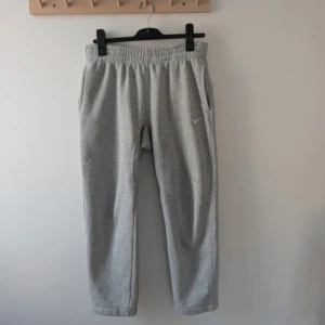 Grå mjukisbyxor från Nike - Grå mjukisbyxor från Nike med elastisk midja och sidofickor. Byxorna har en enkel design med liten Nike-logga på ena benet och en bakficka. Perfekta för en avslappnad stil.