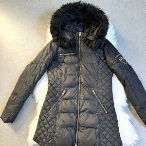 Svart dunjacka med fuskpäls från Rockandbelle - Snygg svart dunjacka från Rockandbelle med fuskpäls på huvan och guldfärgade dragkedjor. Jackan har quiltade partier, tre fickor med dragkedja och en patch på ärmen. Perfekt för kalla dagar.