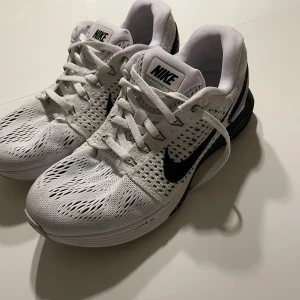 Vita och svarta Nike löparskor - Säljer ett par vita Nike löparskor med svart swoosh och ventilerande mesh-ovandel. Skorna har snörning och en platt sula med stabil häl. Perfekta för träning eller löpning. Snygg och sportig design.