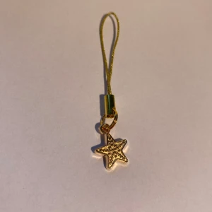 Hänge med en sjöstjärna i guld - Söt accessoar med en liten stjärnformad berlock i guld med små detaljer. Hängs i en tunn, glittrig snodd som passar perfekt till mobil, väska eller nyckelknippa till sommaren